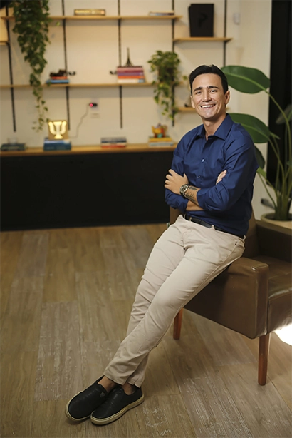 imagem do Pablo sentado no braço de uma cadeira marrom, com um vaso de planta atrás, vestindo uma camisa azul e calça bege, com os braços cruzados sobre o peito, sorrindo